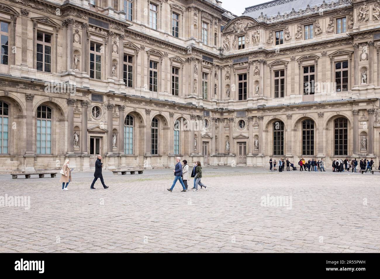 Long lines of visitors in the courtyard of the elegant Palais Royale waiting to enter the Louvre art musuem on a tranquil grey spring day in Paris, France. Amidst a bustling crowd of visitors and tourists, these architectural masterpieces blend modernity and history, inviting exploration and appreciation. Witness the harmonious blend of old and new, as the city's rich cultural heritage comes to life amidst the vibrant energy of a popular travel destination. Stock Photo