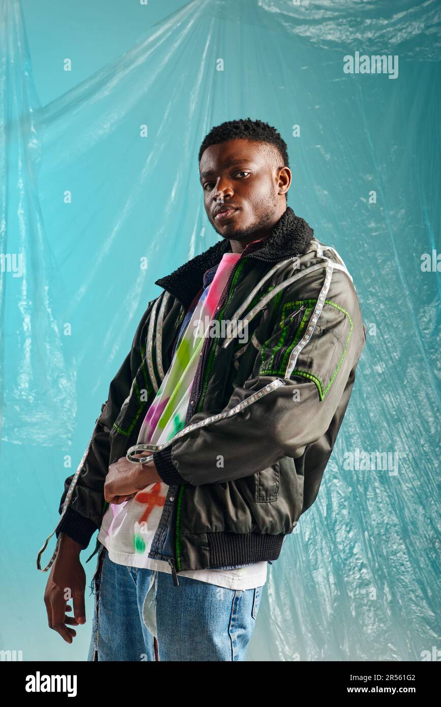 Good looking afroamerican male model in outwear jacket with led stripes looking at camera near cellophane on turquoise background, urban outfit and mo Stock Photo