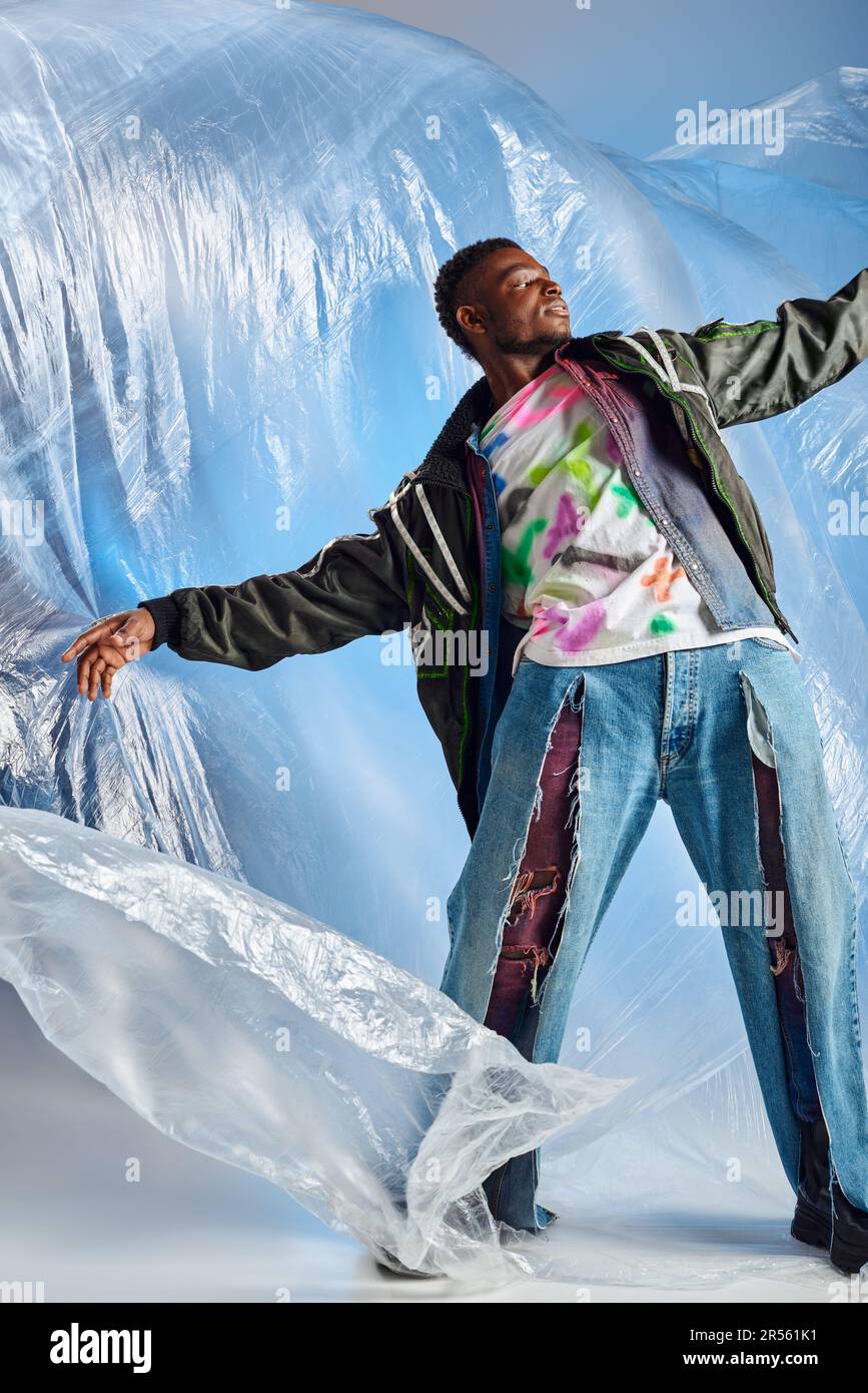 Trendy young afroamerican model posing in outwear jacket with led stripes and ripped jeans while standing on glossy cellophane on blue background, urb Stock Photo