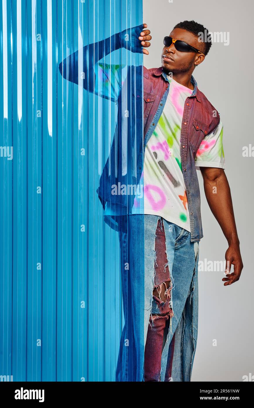 Good looking young afroamerican model in sunglasses, denim vest and t-shirt touching blue polycarbonate sheet and standing on grey background, fashion Stock Photo