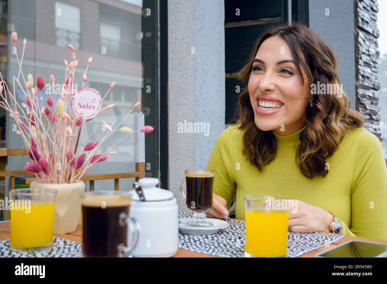 young latin woman of argentinian ethnicity, sitting outside the cafeteria laughing chatting with someone paying attention, while having breakfast and Stock Photo