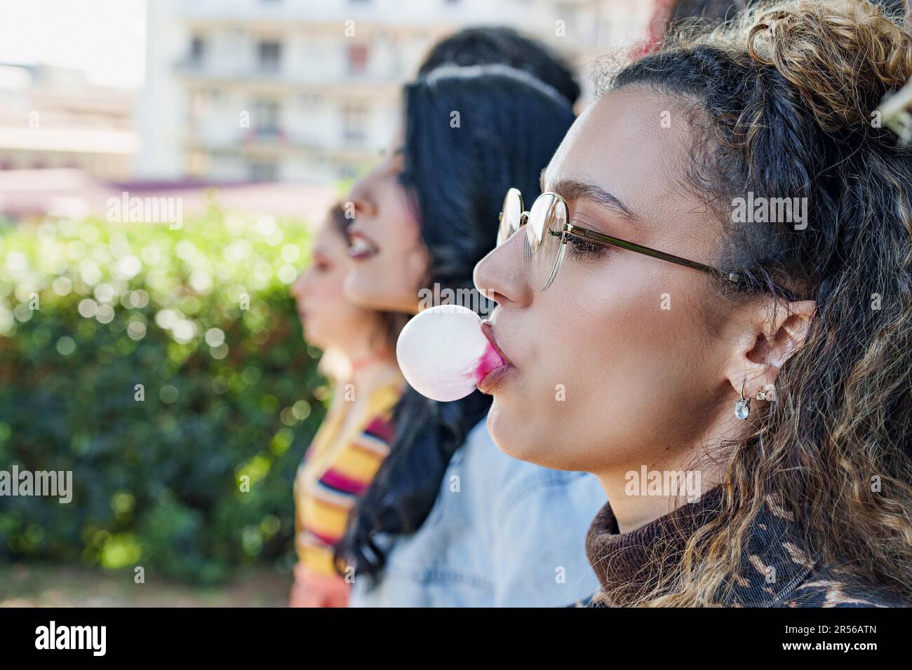 Caucasian woman blowing up a chewing gum balloon in a public park. Playful and carefree atmosphere with friends in the background Stock Photo