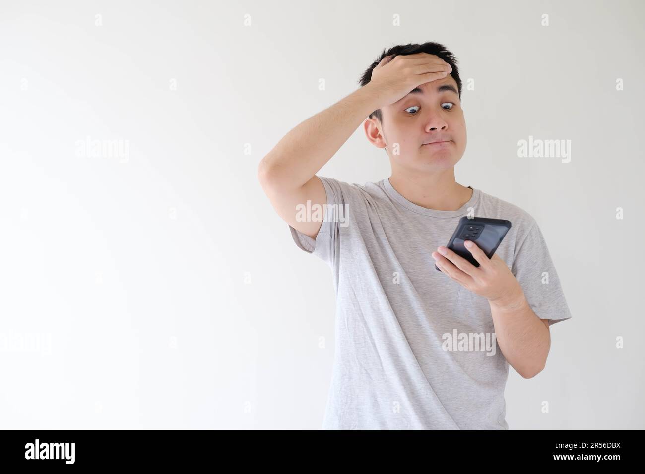 Medium and soft-focused shot of a young Asian man who wears a grey t-shirt and is facepalm while holding while looking at a smartphone. Shallow depth- Stock Photo