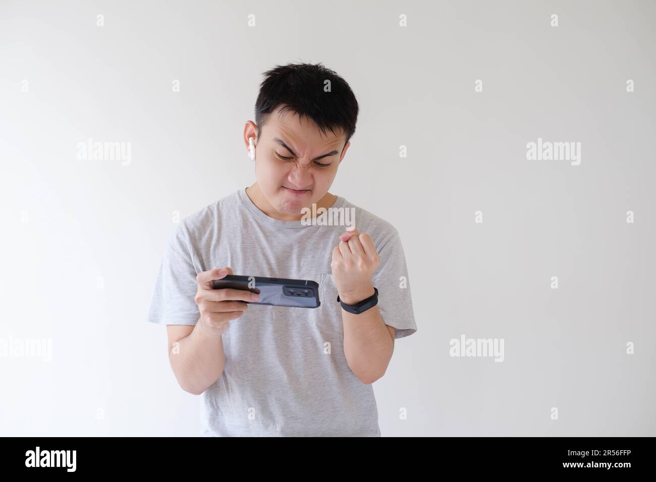 A young Asian man wearing a gray t-shirt is angry while playing a game on his smartphone. Isolated white background. Stock Photo