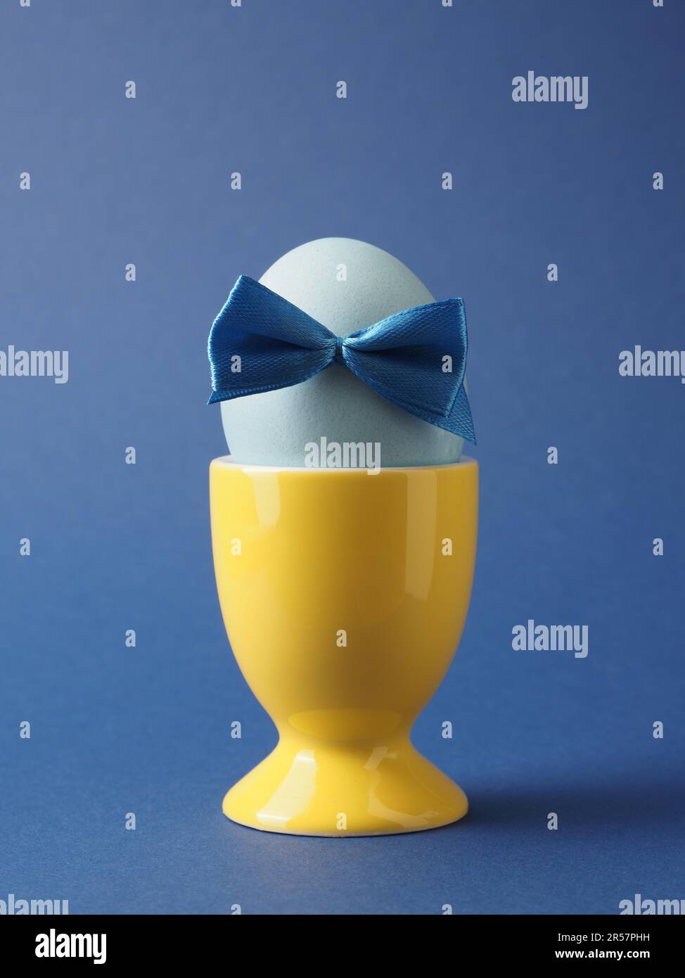 Organic Easter egg with a blue bow in a yellow egg cup on a blue background, happy Easter background Stock Photo