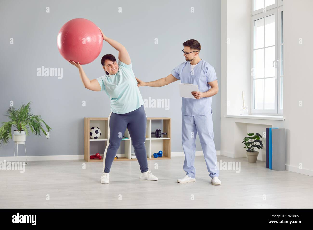 Physiotherapist helping overweight woman patient to do side bends with balance ball Stock Photo