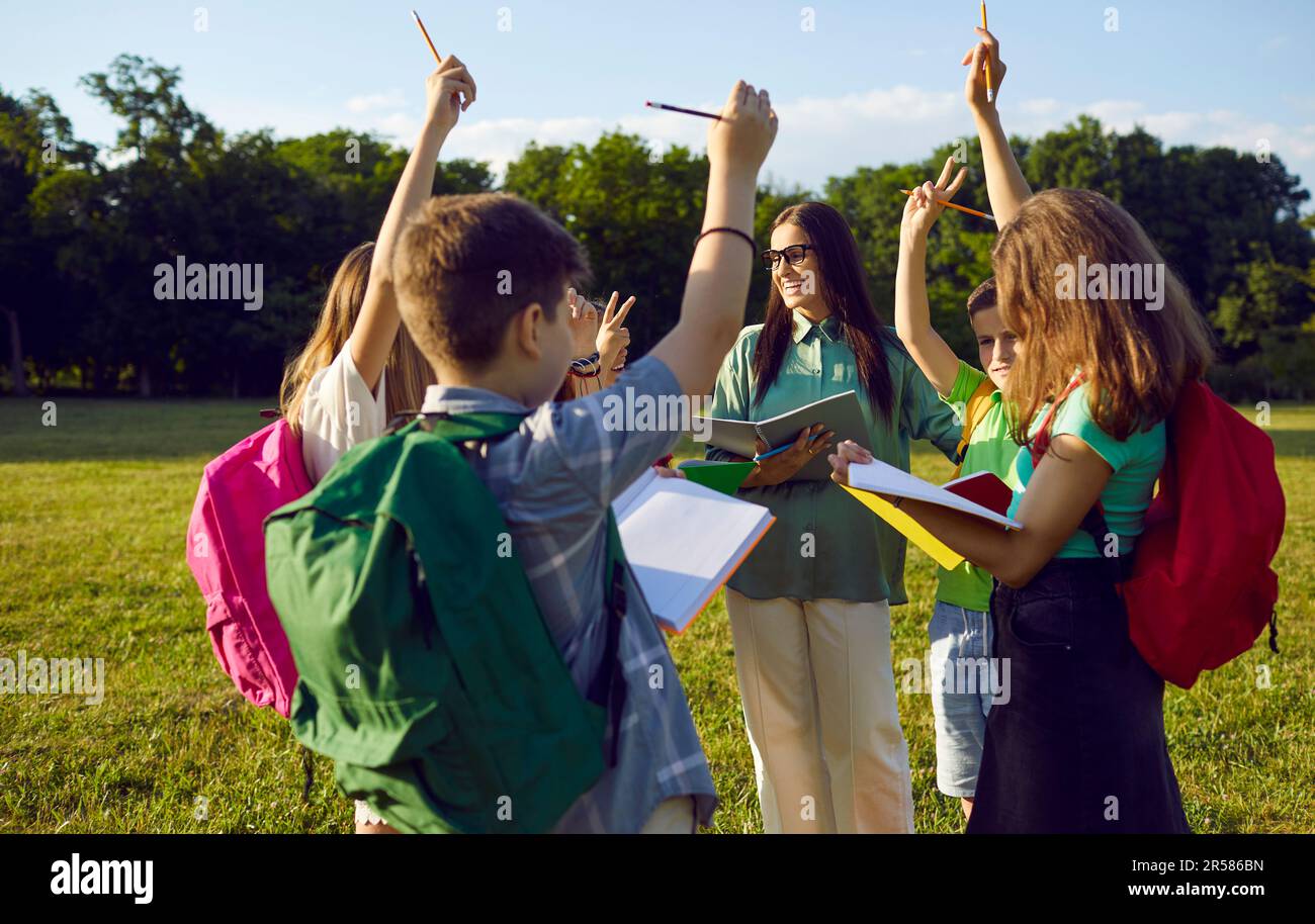 Group of school kids having outdoor lesson and raising hands to answer teacher's question Stock