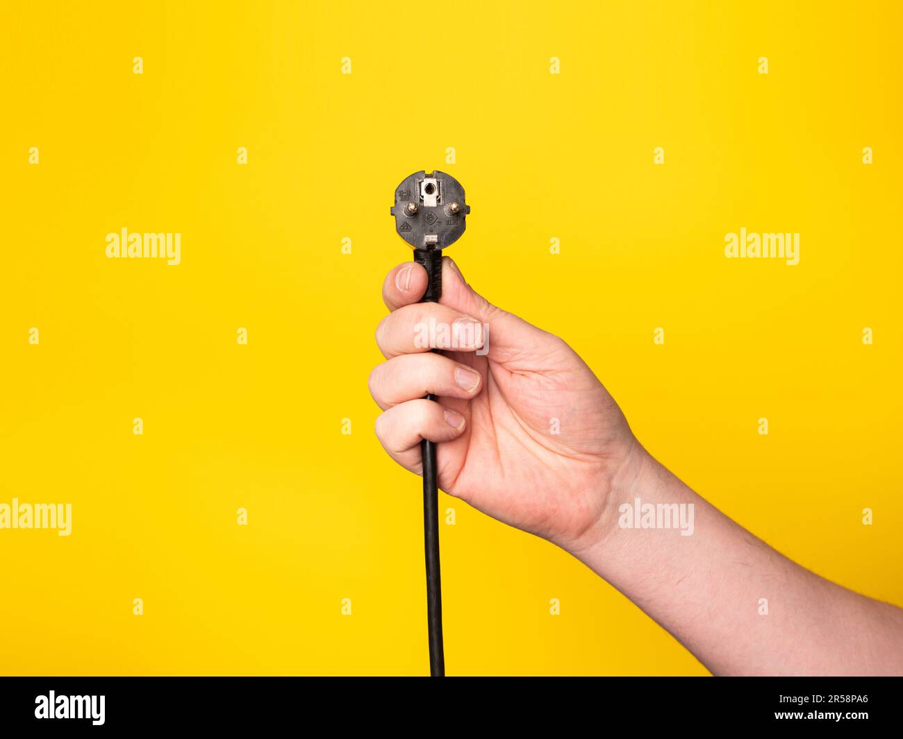 A hand holds a plug to which a black cable is attached.  No face, yellow background. Stock Photo