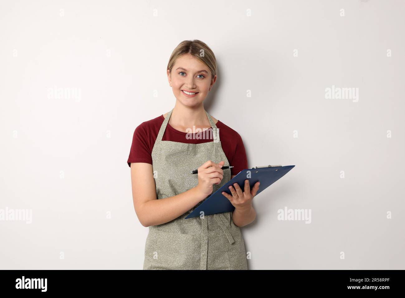 Beautiful young woman in clean apron with clipboard on light grey ...
