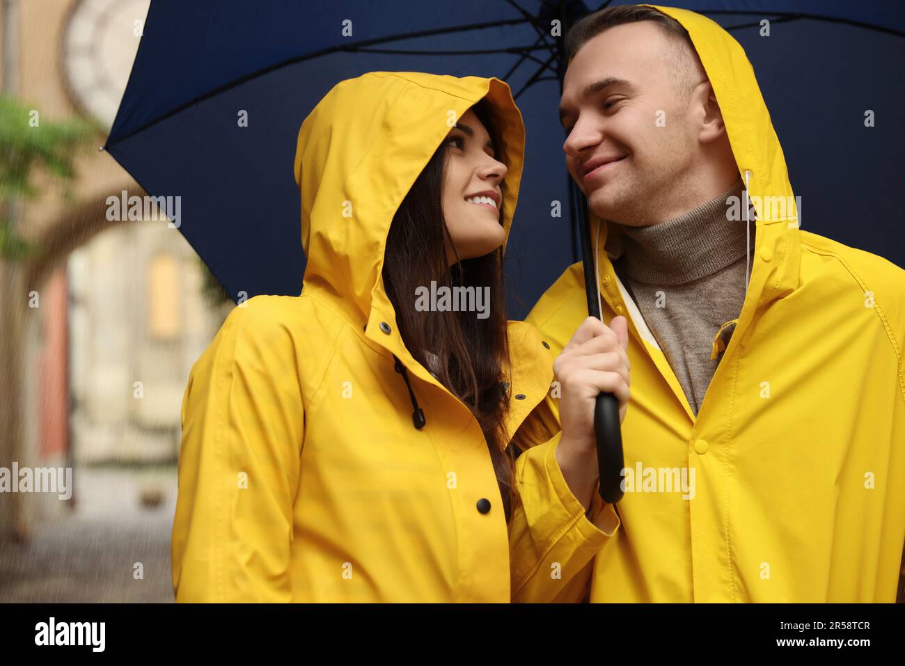 Lovely young couple with umbrella walking under rain on city street ...