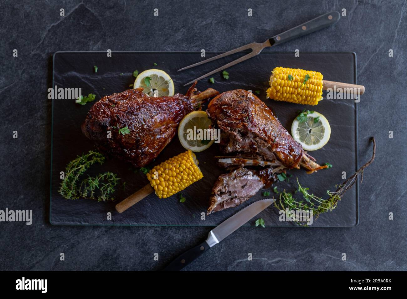 Meat dish with grilled or baked turkey legs. Served with corn on the cop on a platter with garnish on dark background from above Stock Photo