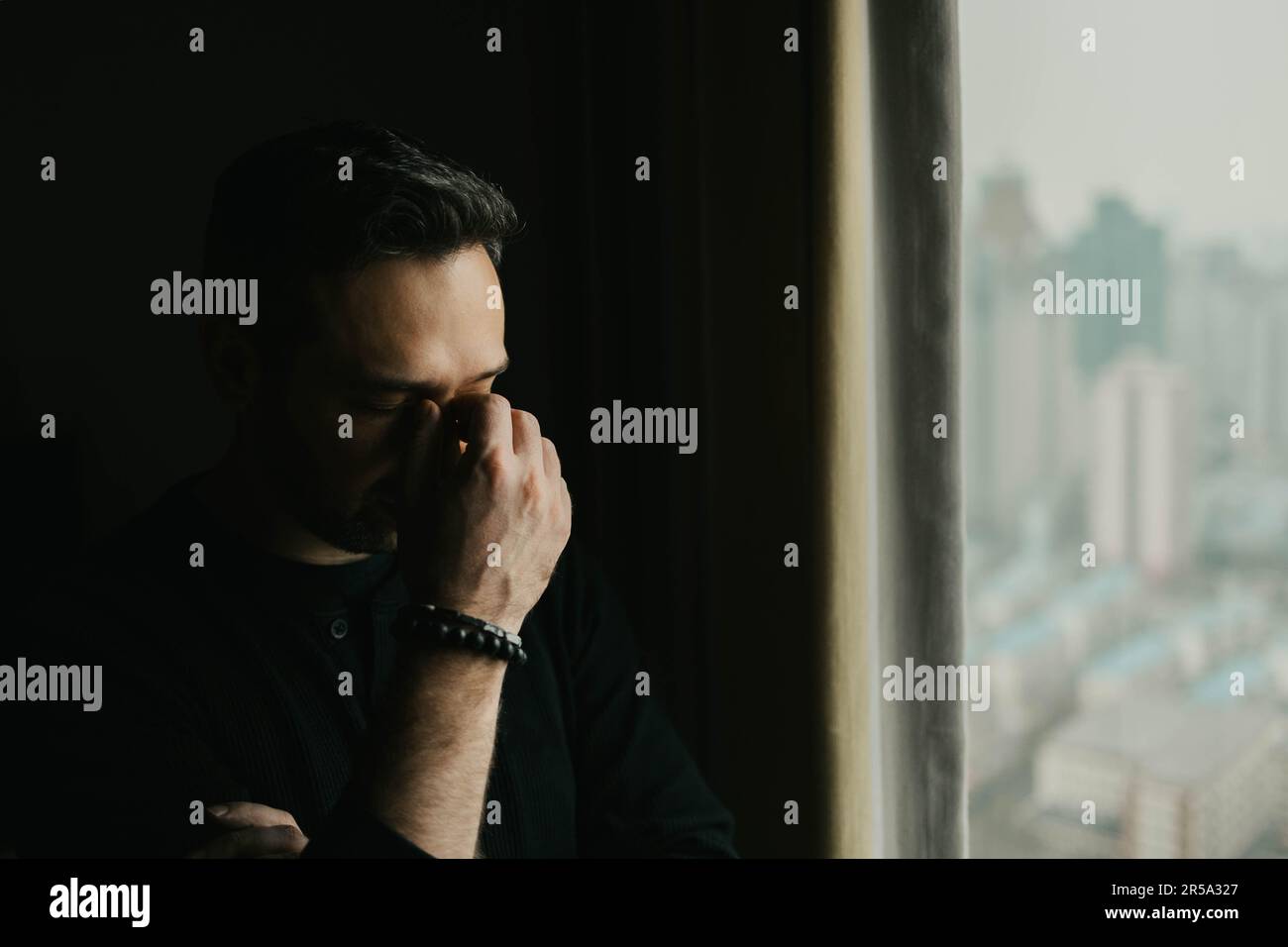Frustrated man stands by a window and puts his hand to his face Stock Photo