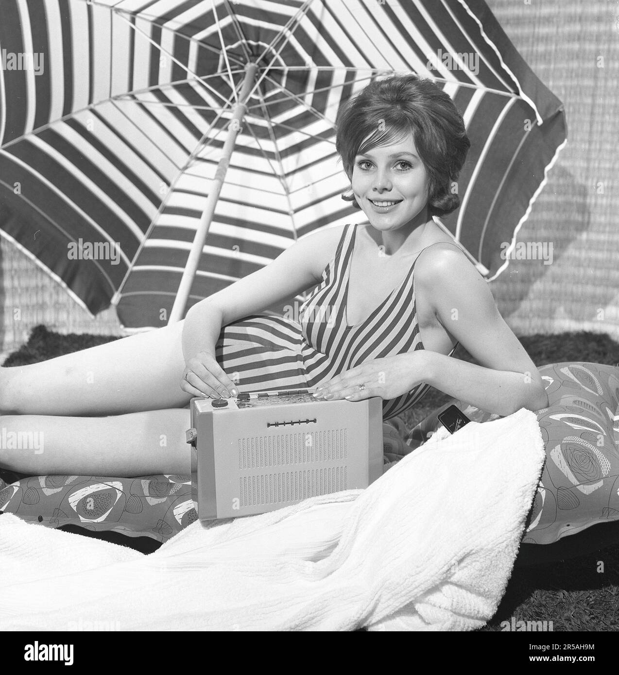 1960s summer lifestyle. A young woman pictured in a photographers studio wearing a bathingsuit with a portable radio. The sunshade parasol in the background is placed there to add to the summer feeling.  Sweden 1960 Photo Kristoffersson Ref CL114-10 Stock Photo