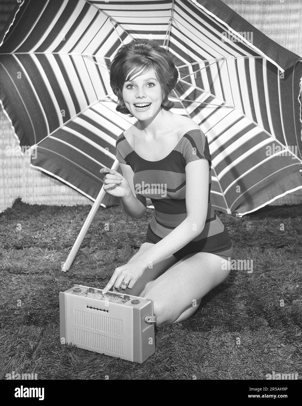 1960s summer lifestyle. A young woman pictured in a photographers studio wearing a bathingsuit with a portable radio. The sunshade parasol in the background is placed there to add to the summer feeling.  Sweden 1960 Photo Kristoffersson Ref CL113-4 Stock Photo
