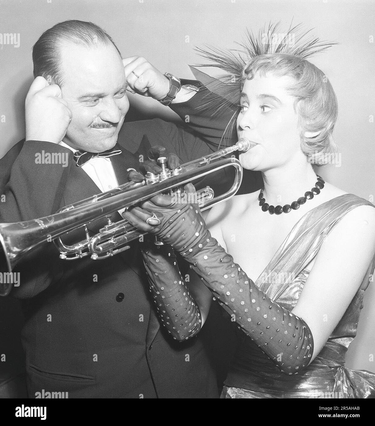 A young woman wearing an evening dress and arm-length gloves plays the trumpet. Judging by the man next to her holding his ears, her musical skills probably leave a lot to be desired. Someone who cannot play the trumpet and who tries to do so usually can only produce noise. Sweden 1955. Kristoffersson BX22-12 Stock Photo