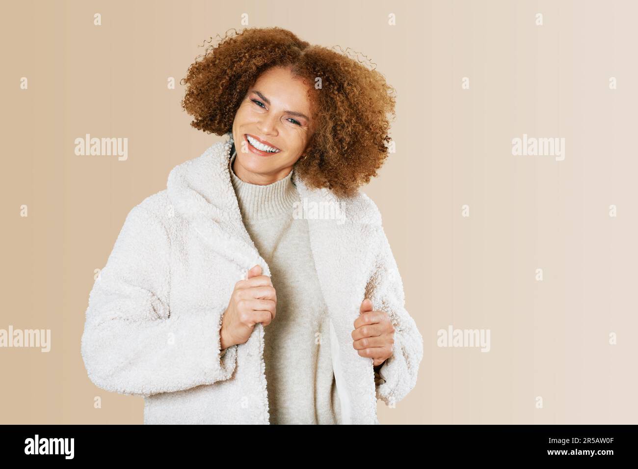 Studio portrait of beautiful middle age woman 50 - 55 years old, wearing natural teddy coat, isolated on beige background Stock Photo
