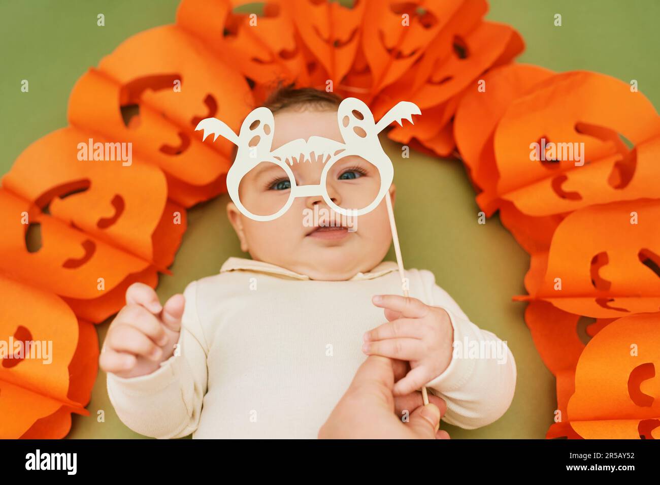 Halloween portrait of adorable baby lying on green background next to pumpkin garland, holding ghost paper glasses for photo booth Stock Photo