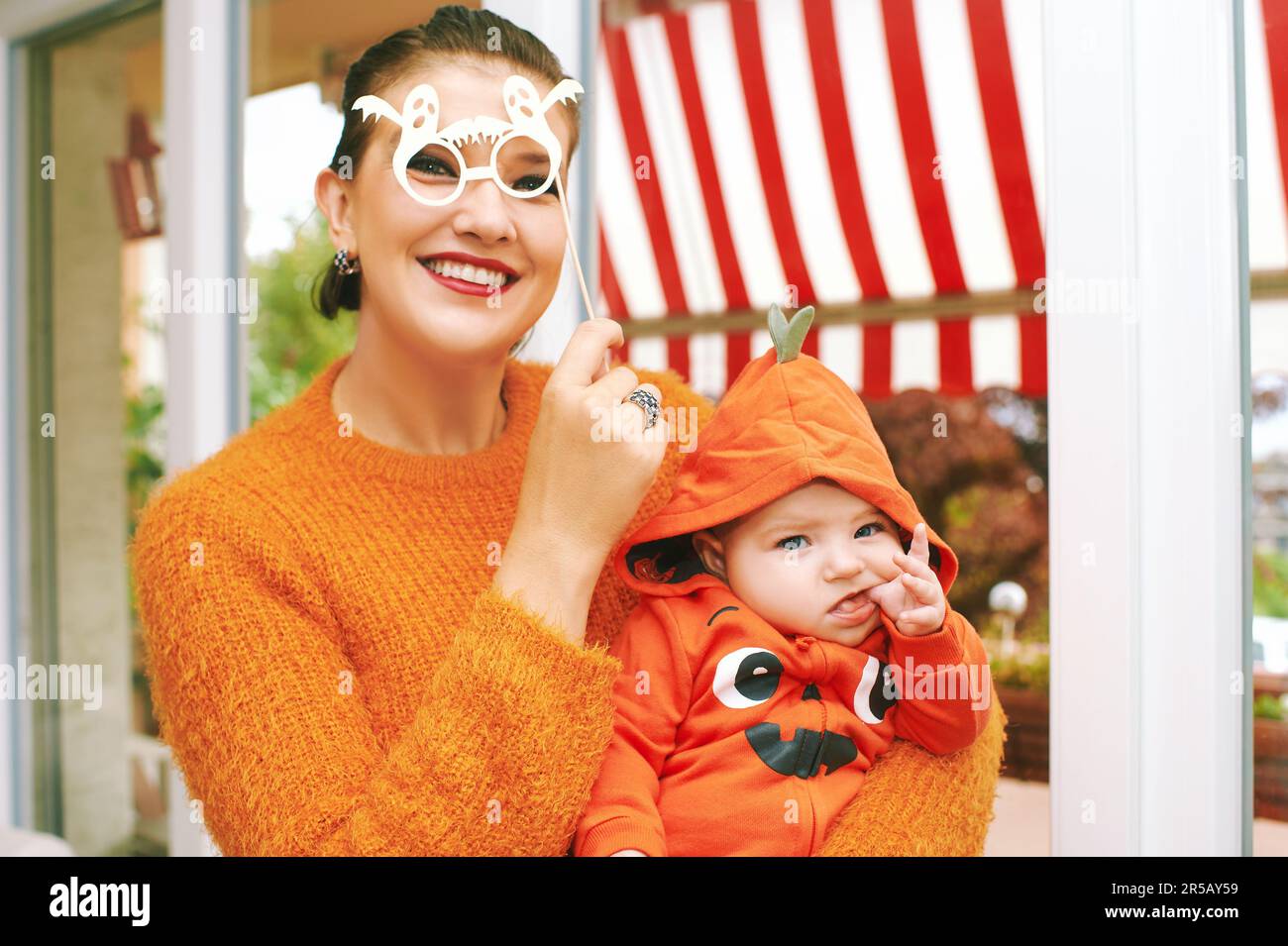 Halloween party with baby, mother holding child wearing pumpkin costume, posing with ghost paper glasses Stock Photo