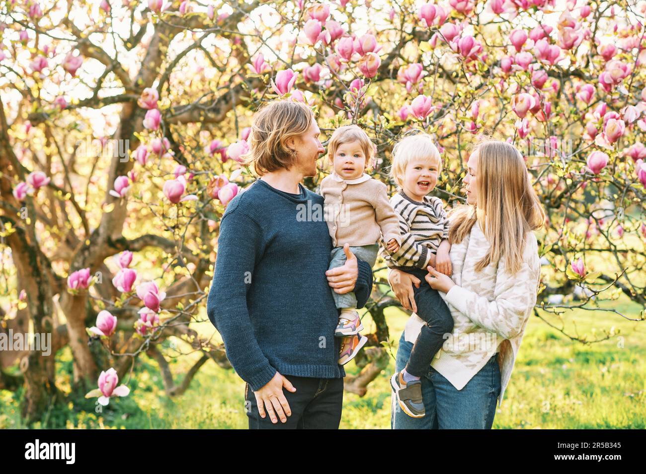 Outdoor portrait of happy young family playing in spring park under blooming magnolia tree, lovely couple with two little children having fun in sunny Stock Photo