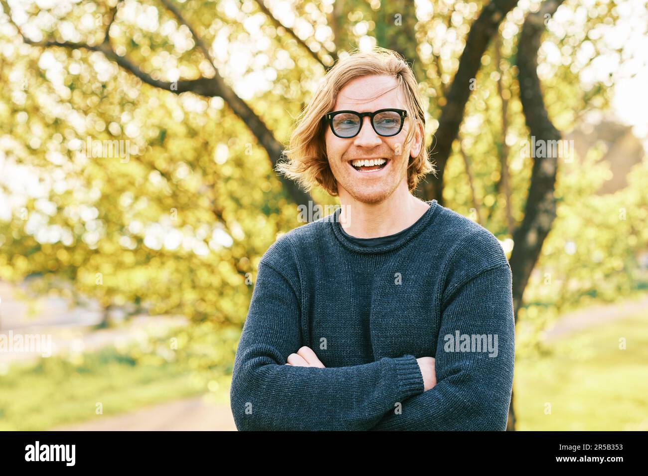 Outdoor portrait of handsome 35 - 40 year old man with red hair, posing in green sunny park, wearing blue pullover and sunglasses Stock Photo