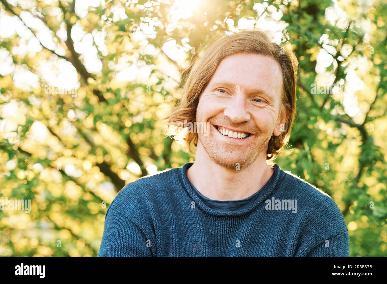 Outdoor portrait of handsome 35 - 40 year old man with red hair, posing in green sunny park, wearing blue pullover Stock Photo