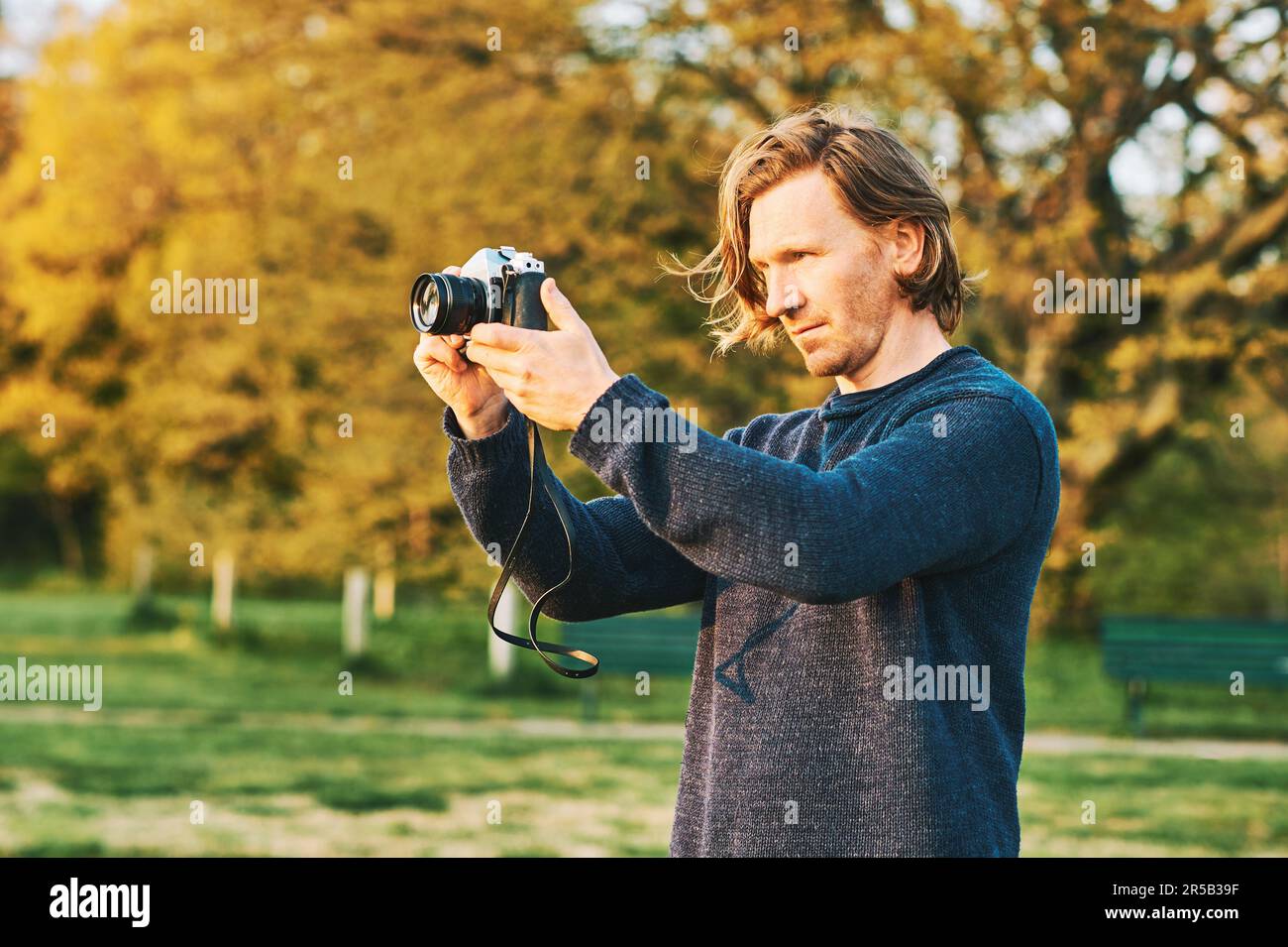 Outdoor portrait of handsome young man taking pictures on green sunny park with old vintage film camera Stock Photo
