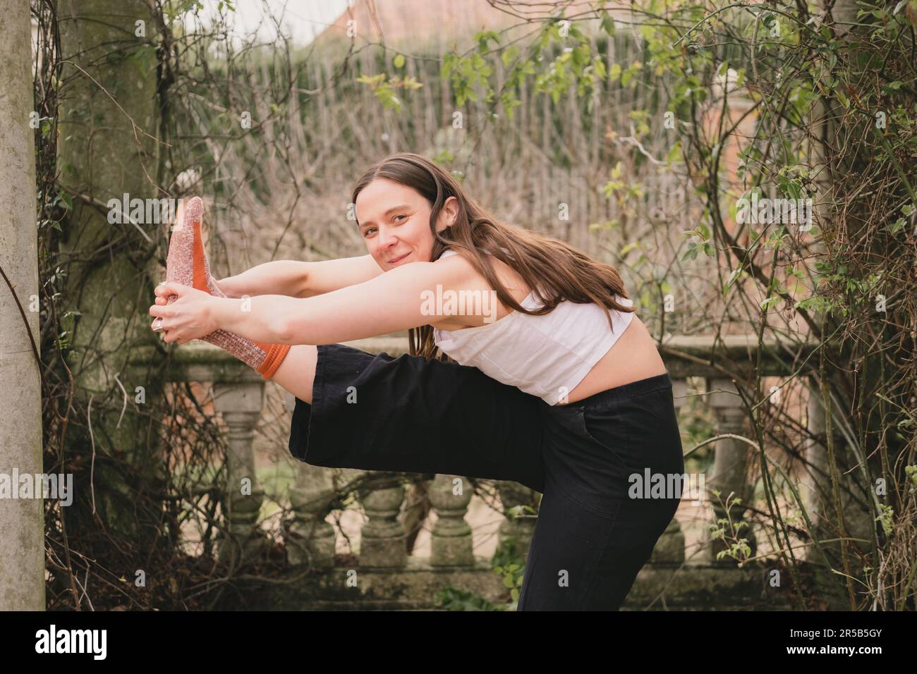 Happy smiley female yoga teacher looking at camera in modified extended hand to toe pose. Wearing black and white loose casual clothes and orange sock Stock Photo