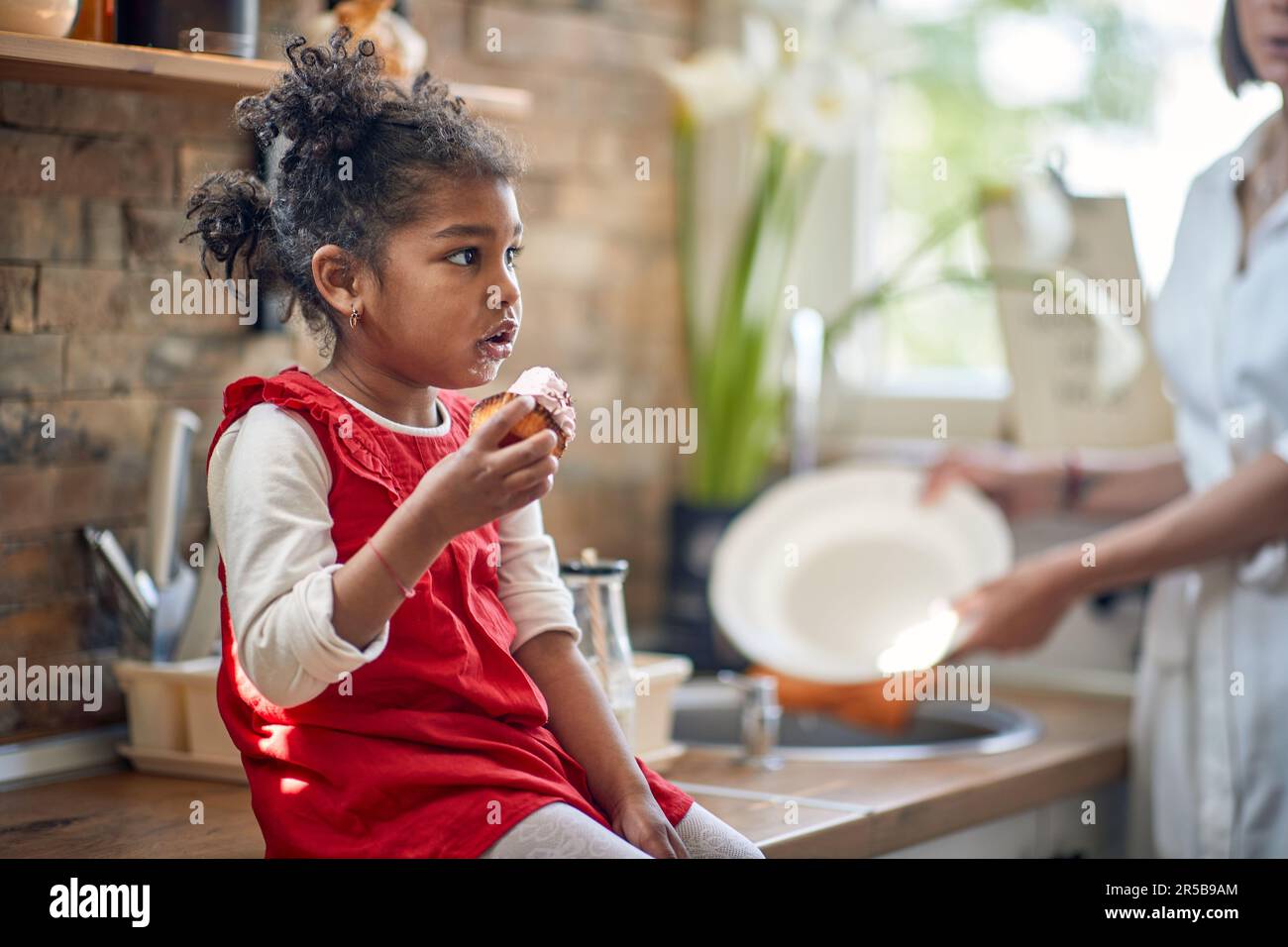 Toddler girl sits in the kitchen, delighting in a mouthwatering muffin, while her mother stands beside her, diligently washing dishes. Stock Photo