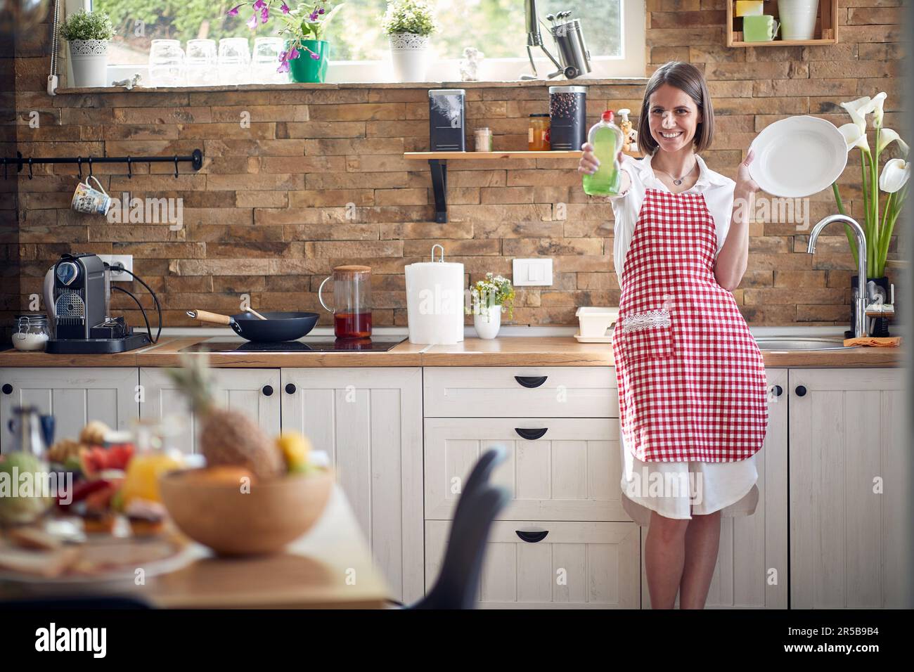 Happy woman stands in her kitchen, radiating joy as she embraces the transformative magic of our exceptional dish soap. Stock Photo