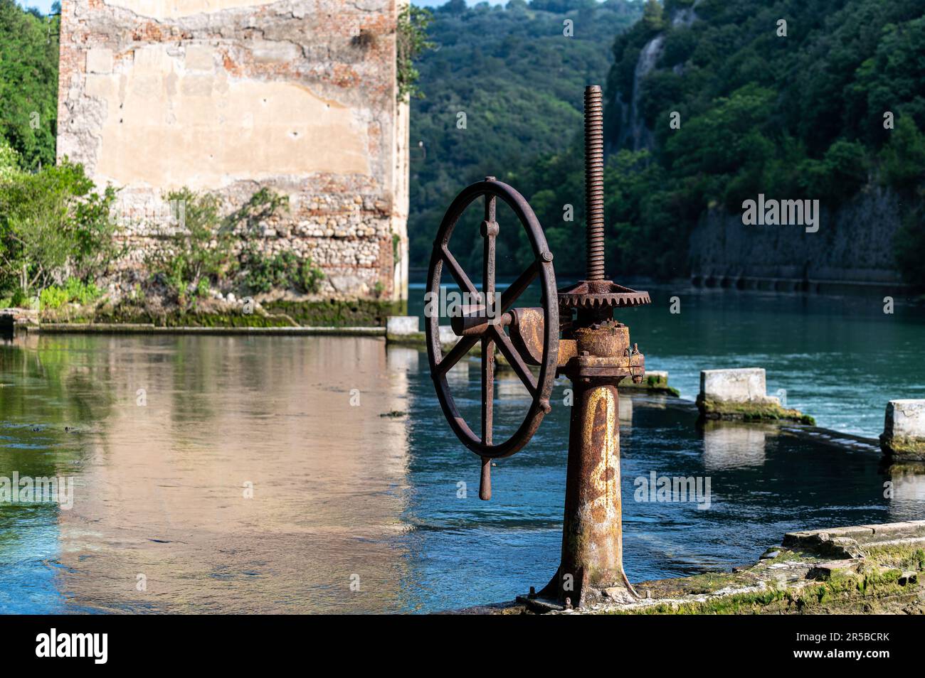 This image features a traditional well situated close to a river, with a wooden water wheel that is visibly rotating Stock Photo