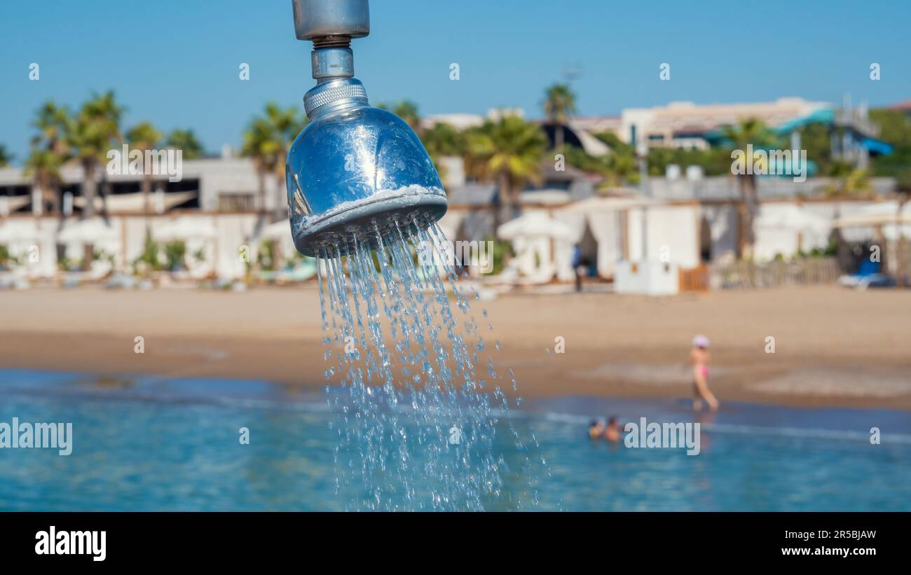 Outdoor refreshing shower head  spraying water to take shower on the pier. Blurry beach and swimming people in the background. Stock Photo