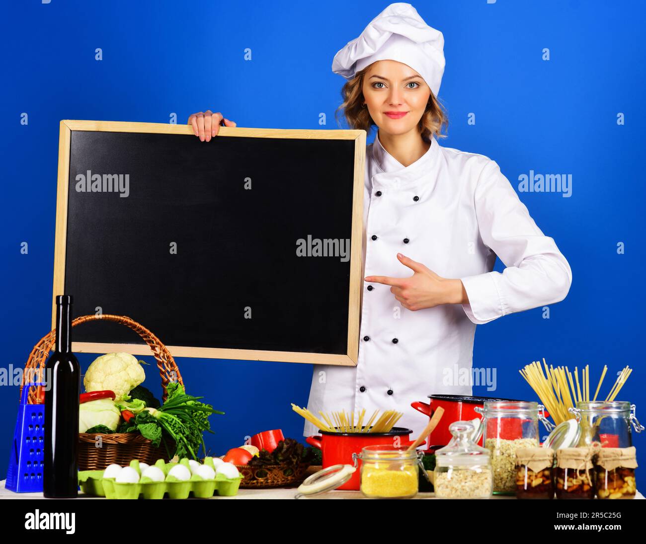 Woman chef pointing finger to blank blackboard. Food preparation. Female cook in chef uniform with empty billboard for advertising text menu in restau Stock Photo
