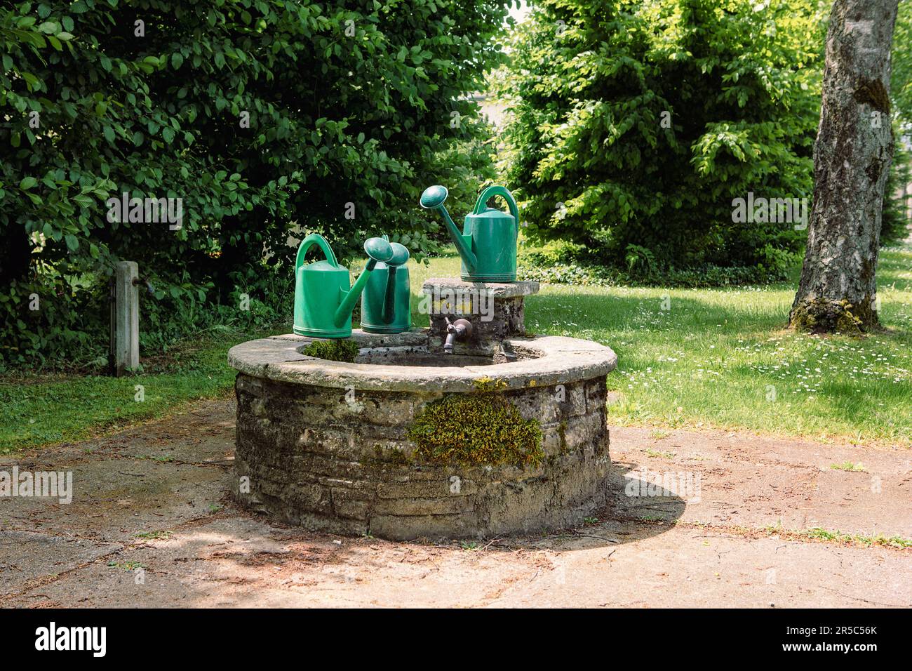Group of green garden watering can in front of stone well. Romantic garden or parc, summertime secret garden scene. Stock Photo