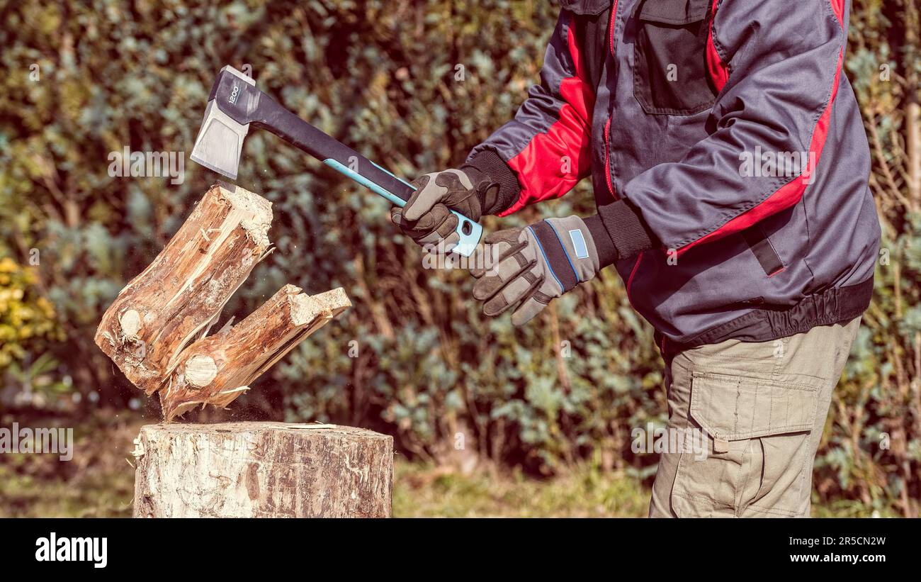 Working woodcutter holding splitting axe in hand and chopping wood log on wooden block. Firewood preparation using modern steel ax tool. Energy crisis. Stock Photo