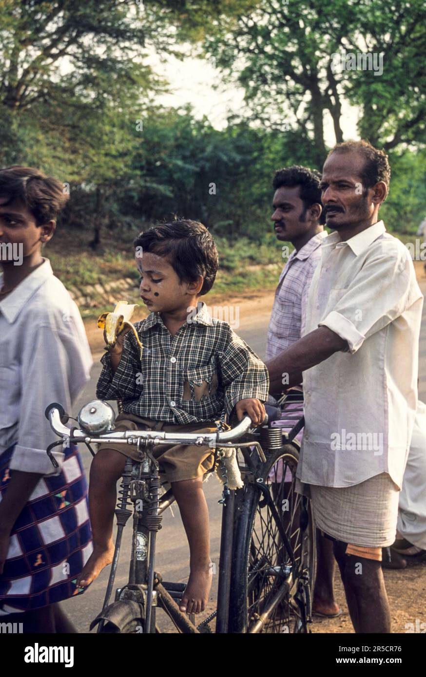 A boy sitting on a bicycle and eating a banana, with his father standing nearby at Avaniyapuram, Madurai, Tamil Nadu, South India, India, Asia Stock Photo