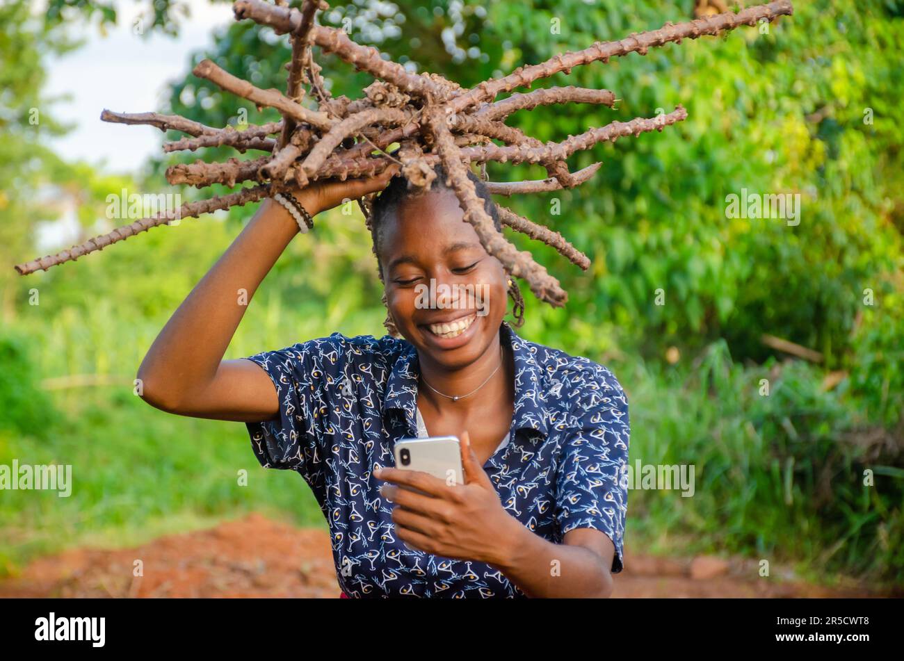 excited young African girl carrying wood and a and using her phone on a farm Stock Photo