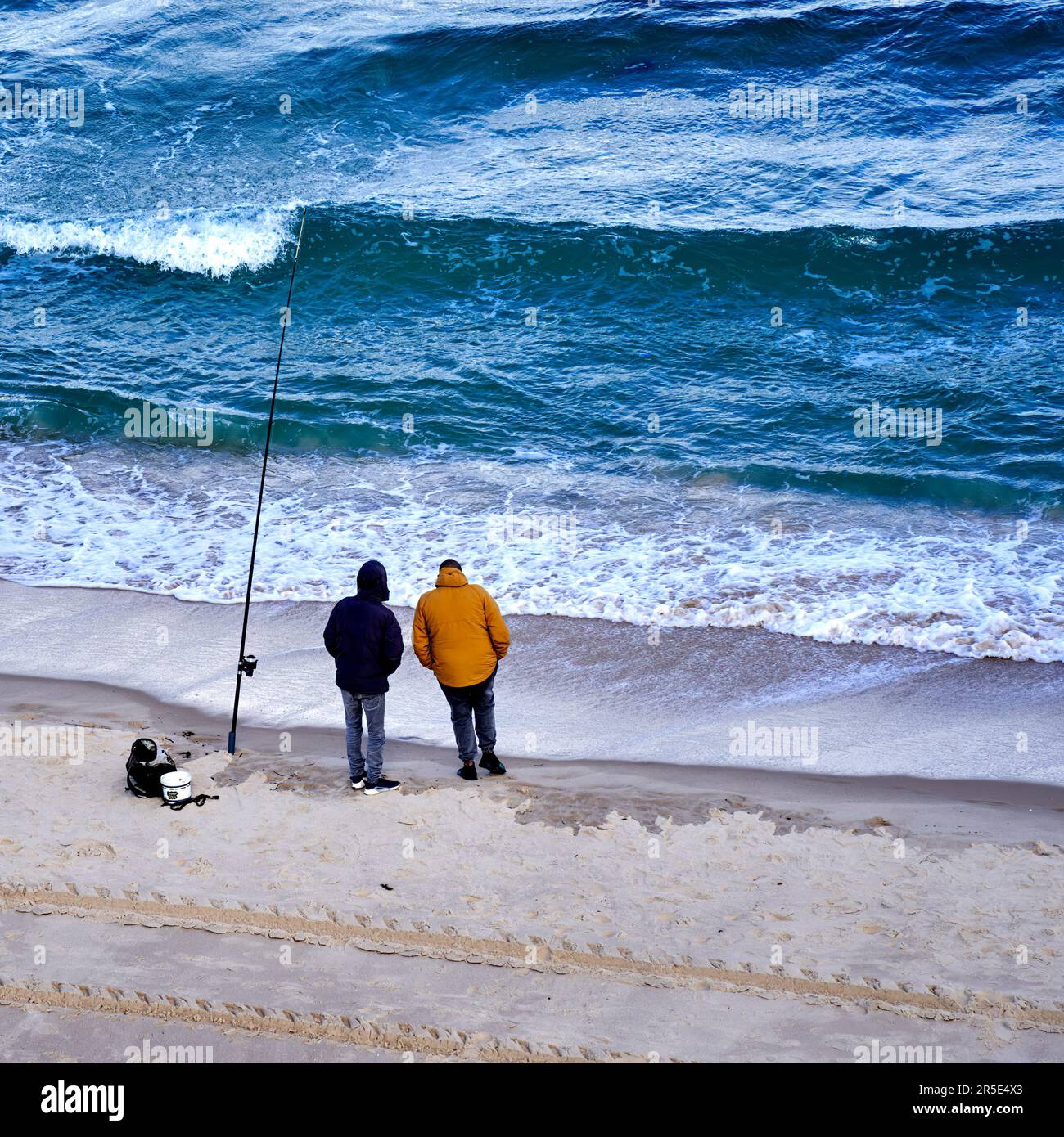 Sousse, Tunisia, January 22, 2023: Angler in warm winter clothes with attached hoods and hands in pockets hold the fishing rod in the Mediterranean Se Stock Photo
