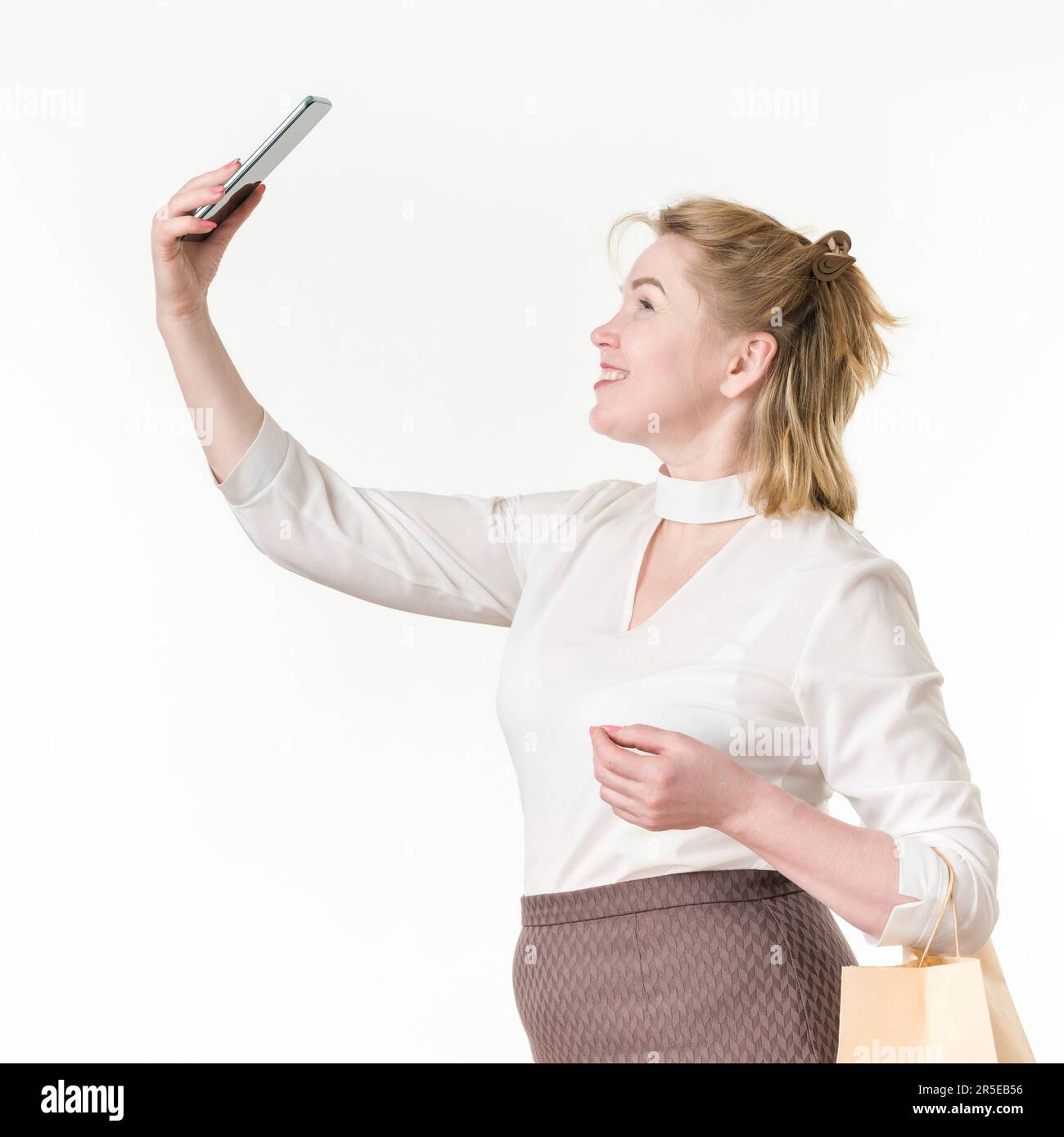 Caucasian ethnicity woman taking selfie holding phone up high, holding shopping bags with purchases. 49 years old smiling woman shopper Stock Photo