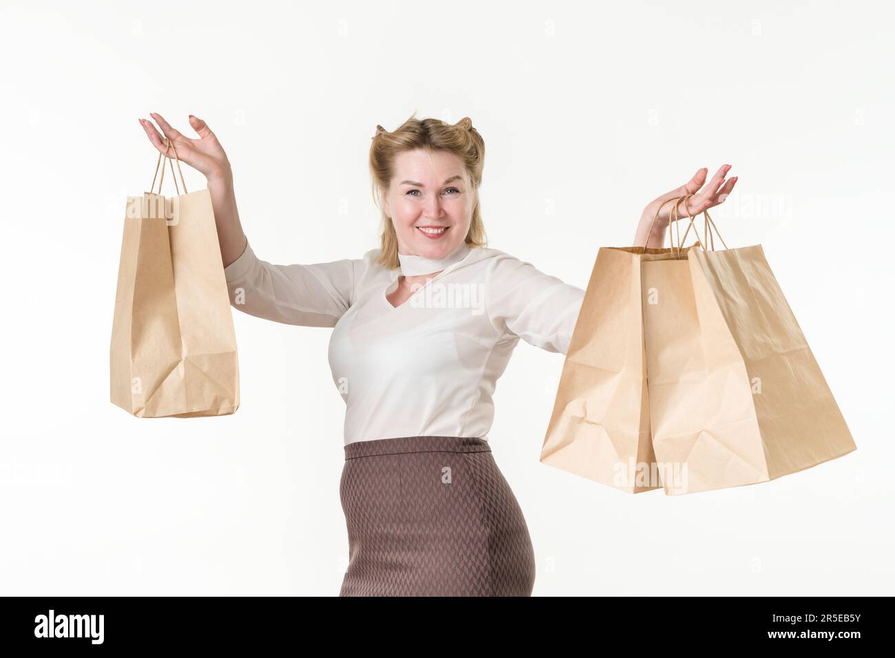 Waist up photo of happy smiling blonde woman holding shopping paper disposable bags with purchases. Female model 49 years old dressed in white blouse Stock Photo