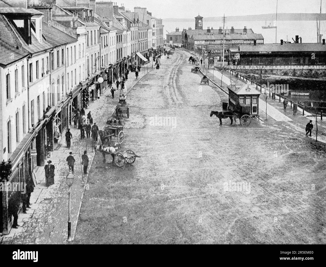 A late 19th century view of the High Road, a shop lined street and waterfront in Cobh, known from 1849 until 1920 as Queenstown, a seaport on the south side of Great Island in Cork Harbour in County Cork, Ireland. A long dedicated cruise terminal, Cobh was the departure point for 2.5 million of the six million Irish people who emigrated to North America between 1848 and 1950. Stock Photo