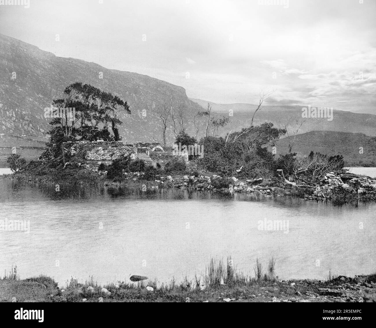 A late 19th century view of the hermitage ruins at Gougane Barra dating from around 1700 when a priest called Denis O'Mahony retreated to the island on a scenic valley lake in the Shehy Mountains of County Cork, Ireland. The name Guagán Barra comes from Saint Finnbarr (or Barra), who is said to have built a monastery on the island in the lake during the sixth century. Stock Photo