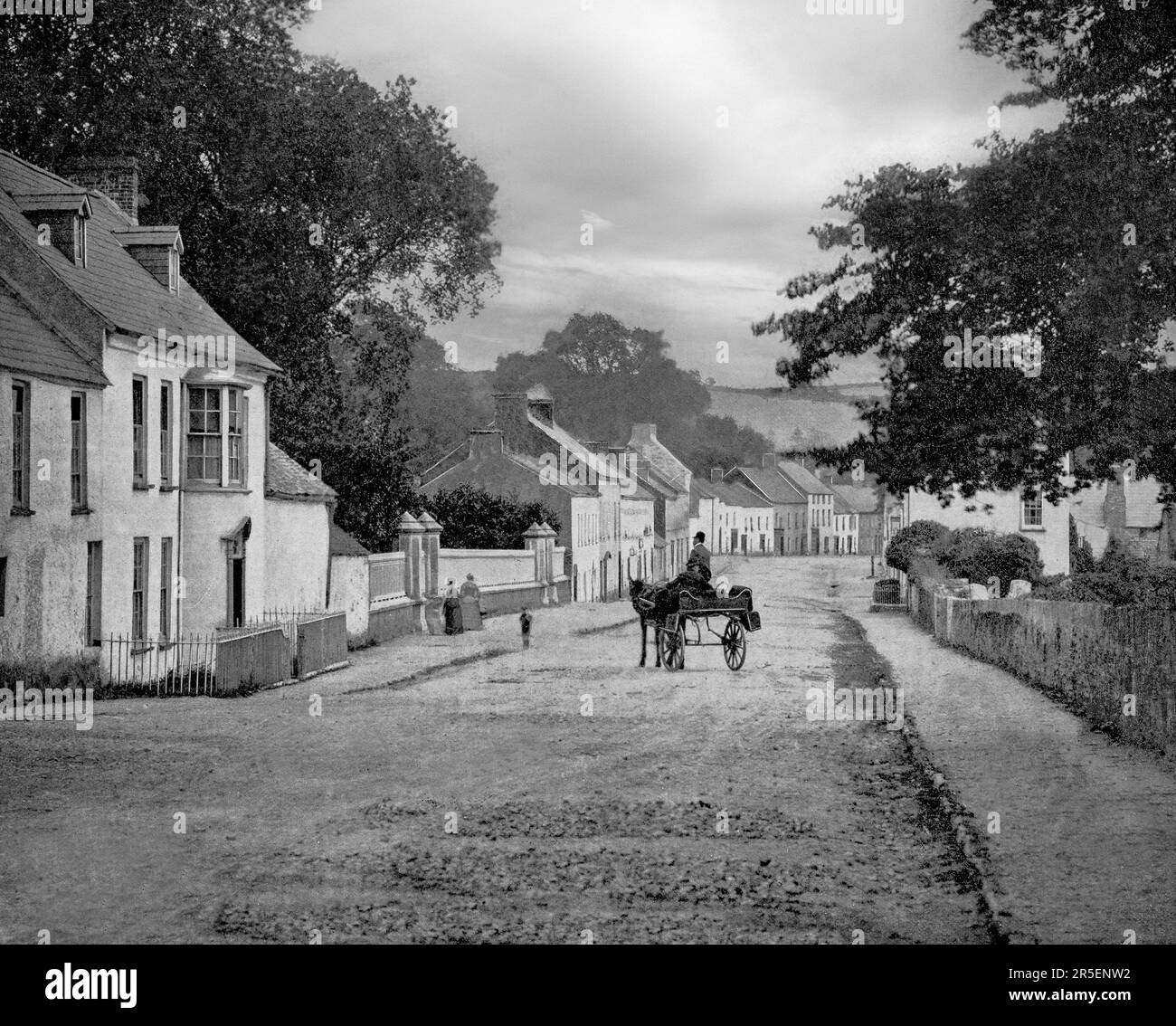 A late 19th century view of a jaunting car in the main street of Inishannon, then little more than a hamlet. Now it is a large village on the main Cork–Bandon road (N71) in County Cork, Ireland. Situated on the River Bandon, the village has since grown due to its proximity to Cork city, and is now a dormitory town for city workers. Stock Photo