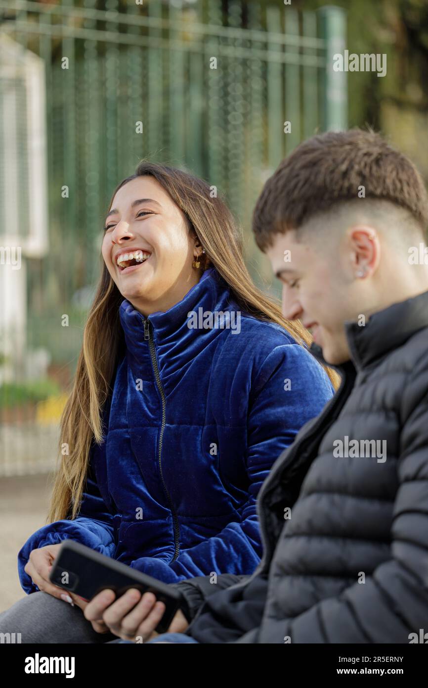 Latina girl laughing sitting on a plaza bench with her boyfriend. Stock Photo