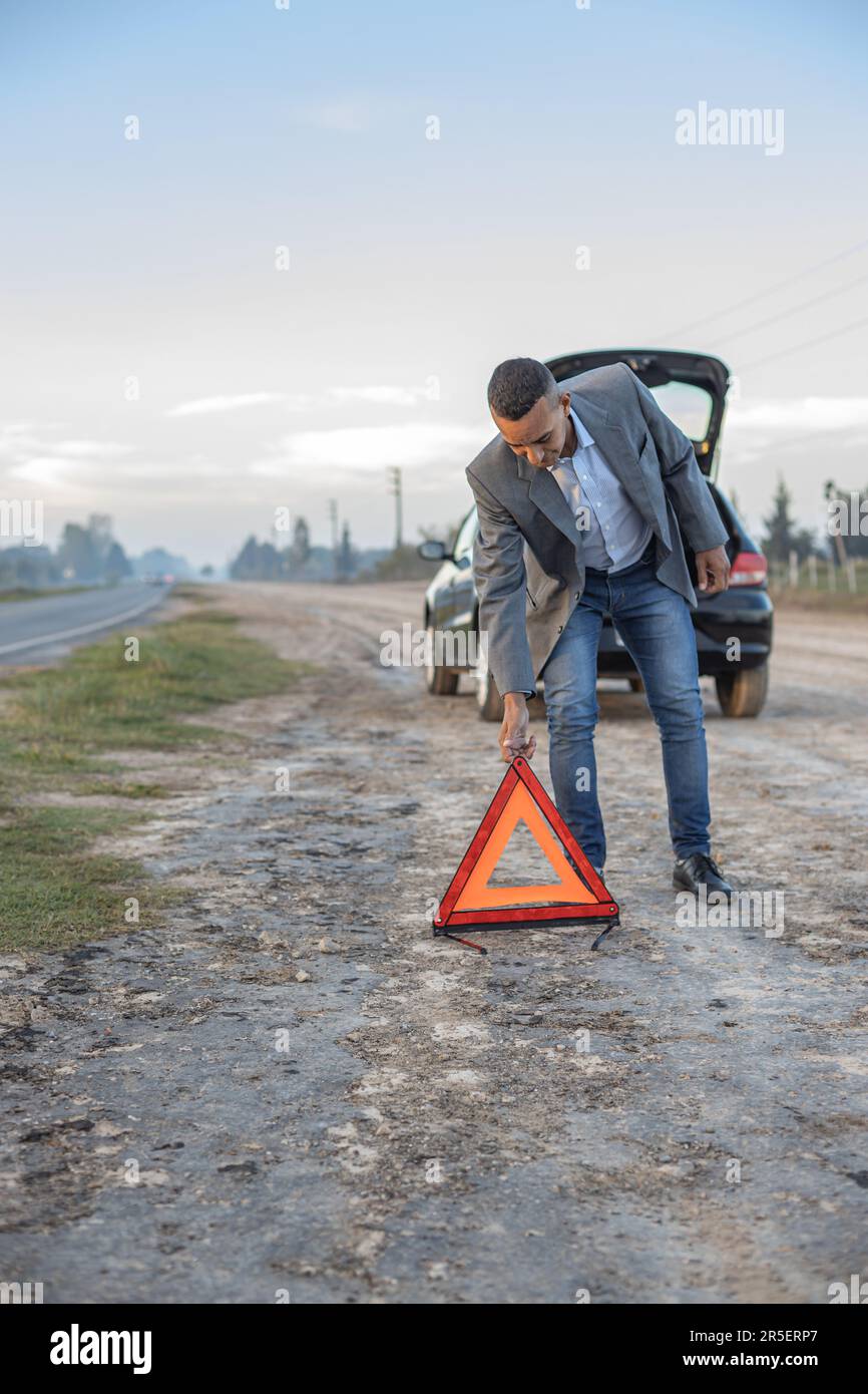 Young Latino man placing a warning triangle behind a broken down car on the side of the road. Stock Photo