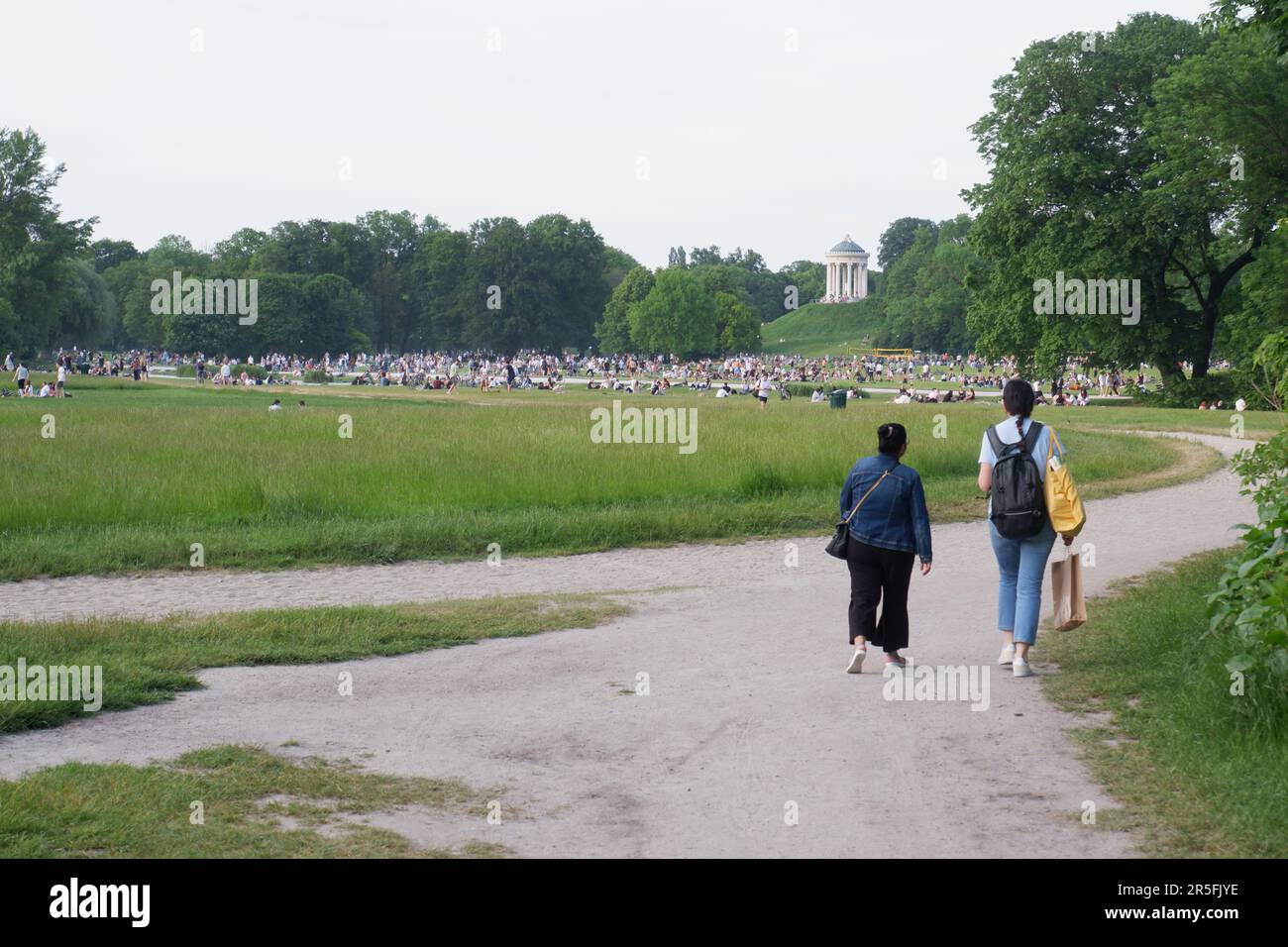 A pair of friends in foreground walk on a path in the Englischer Garten Munich Germany on a June evening, there are large crowds in the park beyond Stock Photo