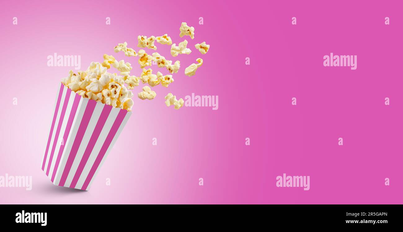 Popcorn flying out of pink white striped paper box isolated on pink background with copy space. Splash, levitation of popcorn grains. Stock Photo