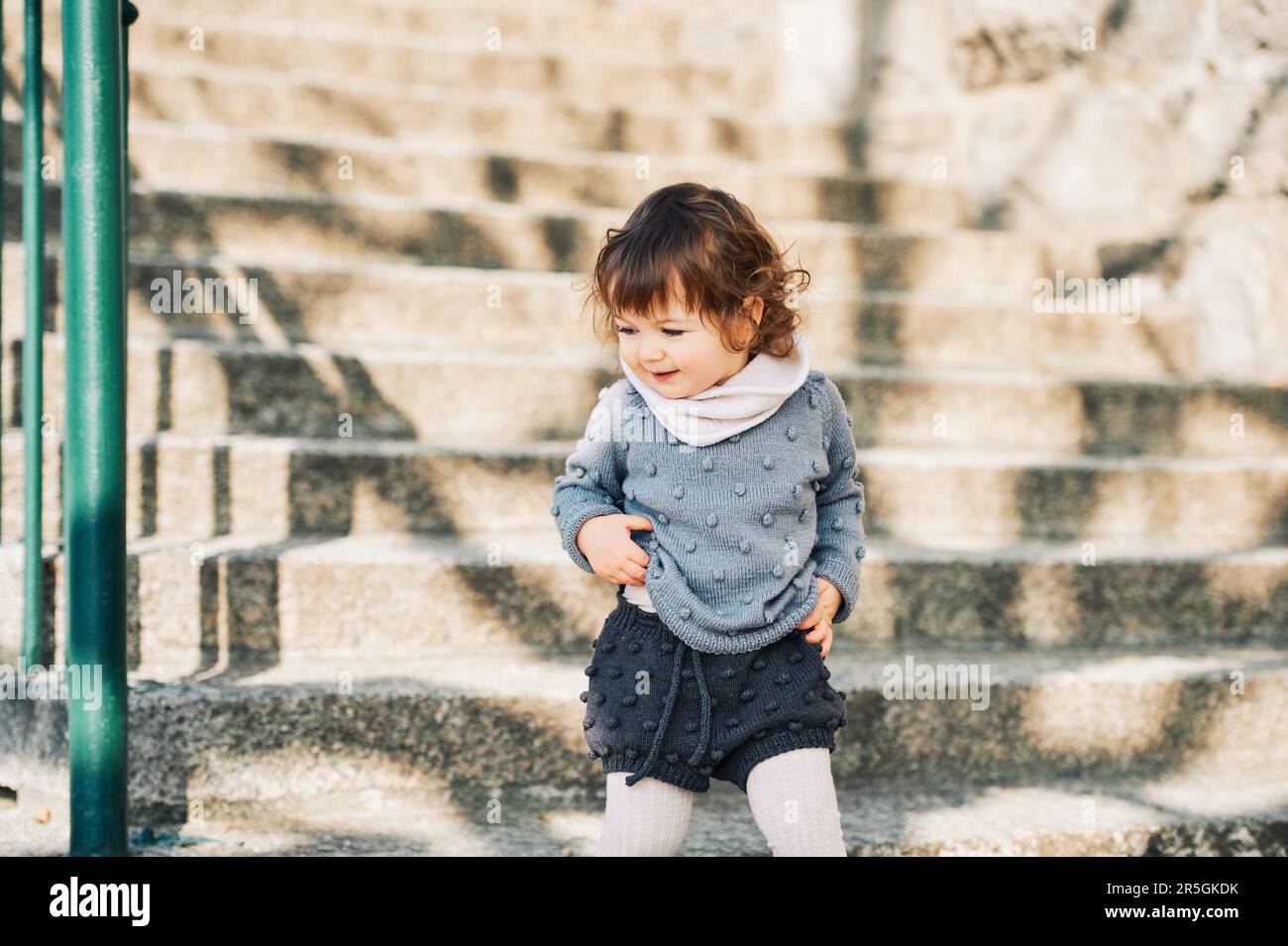 Outdoor portrait of adorable toddler girl walking up the stairs, child of 1 - 2 years old climbing steps on city street, wearing knitted set of pullov Stock Photo