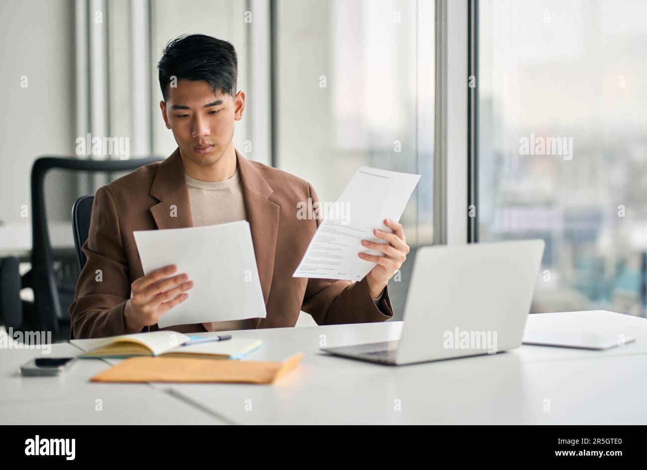 Young busy Asian business man holding documents sitting at desk in office. Stock Photo