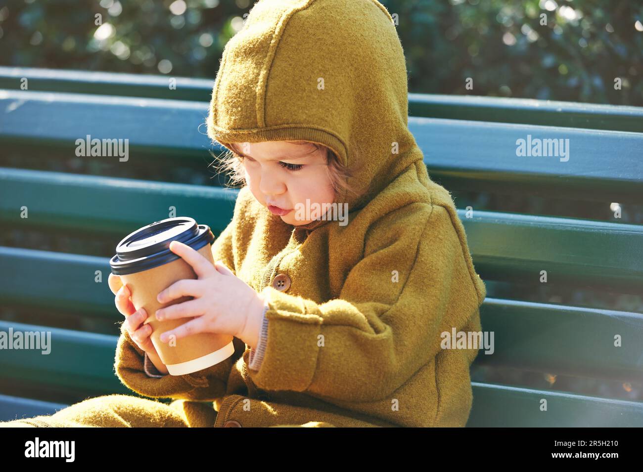 Funny toddler kid resting on bench, holding paper cup of take away coffee Stock Photo