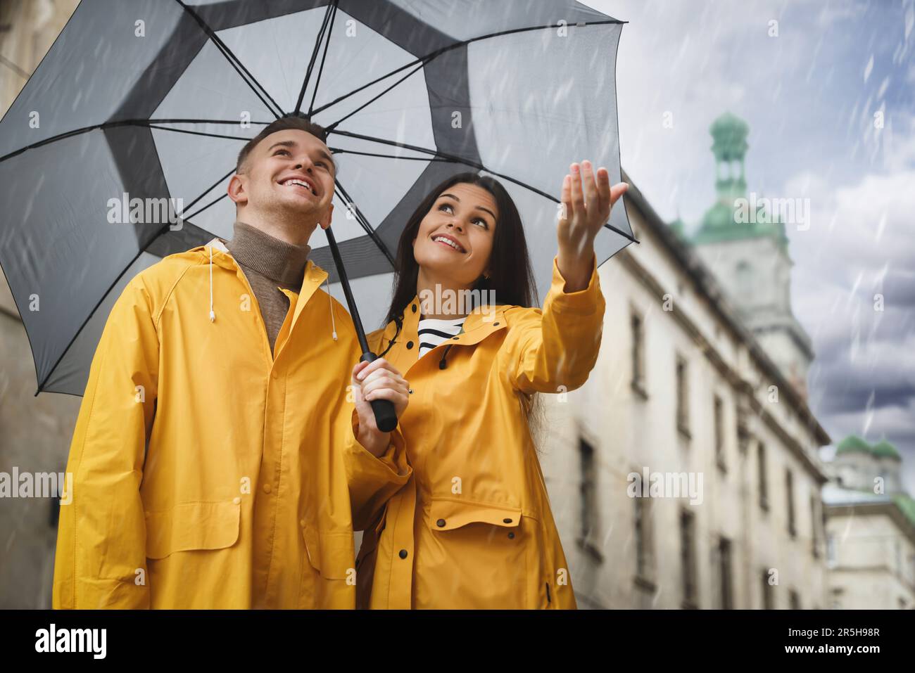 Lovely young couple with umbrella walking under rain on city street ...